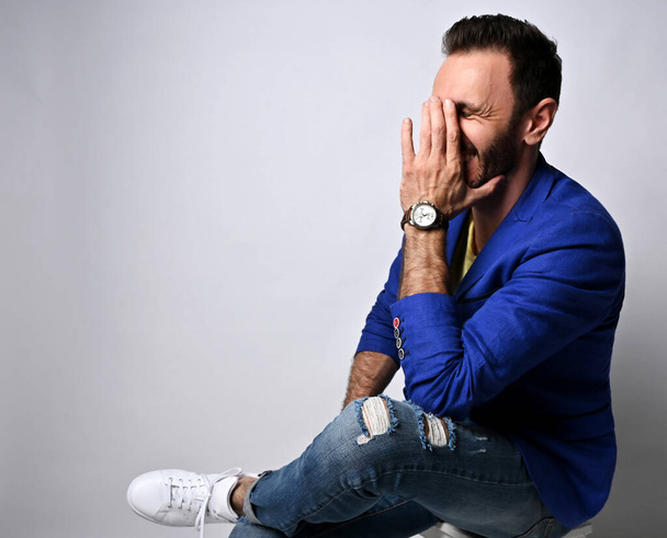 Portrait of adult man in modern clothing jeans and jacket sitting sideways on stool laughing covering face with palm - Фото, зображення