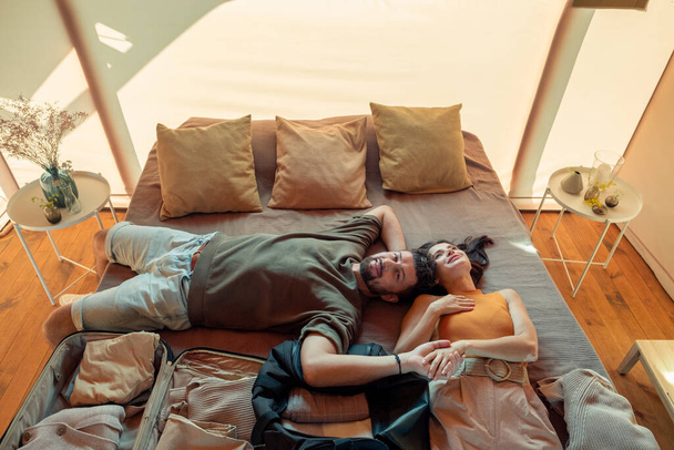 Счастливая пара мечтает лежа на кровати с распакованными сумками - Фото, изображение