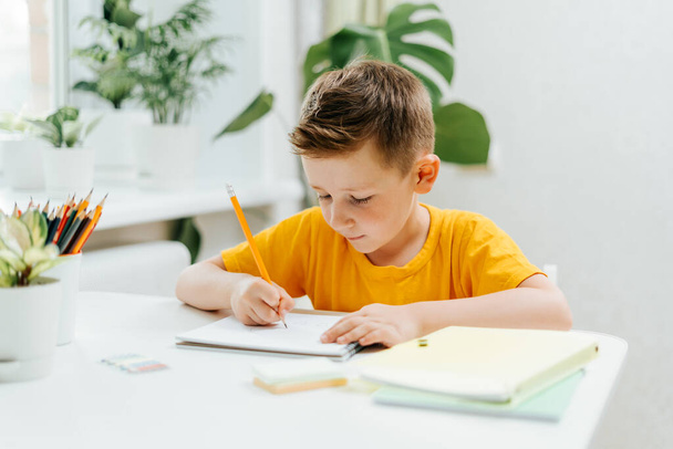 遠隔オンライン教育。本を持って家で勉強している白人の笑顔の子供は、ノートパッドで書き、学校の宿題をしています。ノートと一緒にテーブルに座っている子供を考える。学校に戻る - 写真・画像