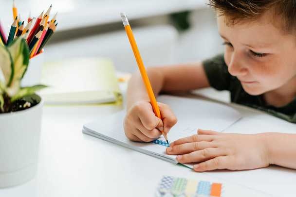 Дистанционное обучение онлайн. Белый улыбчивый мальчик учится дома с книгой, пишет в блокноте и делает домашнее задание. Мыслящий ребенок сидит за столом с блокнотом. Возвращение в школу - Фото, изображение