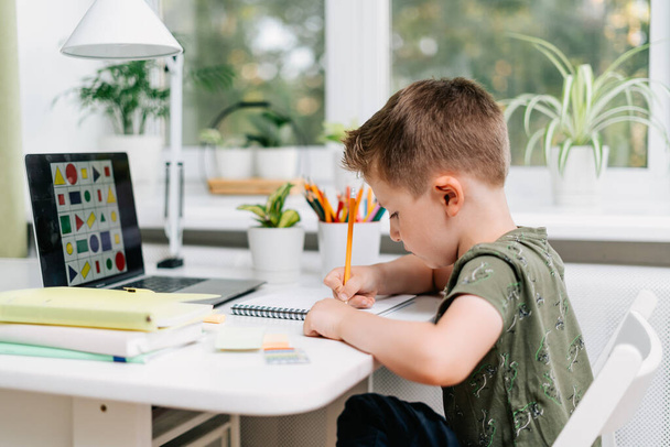 Дистанционное обучение онлайн. Белый улыбчивый мальчик учится дома с ноутбуком и делает домашнее задание. Мыслящий ребенок сидит с блокнотом, карандашами и учебниками. Возвращение в школу - Фото, изображение