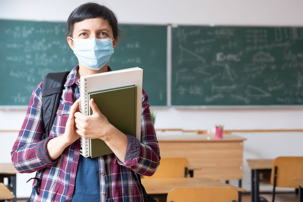 Smiling student girl wearing school backpack, mask and holding exercise book. Social distanting and classroom safety during coronavirus epidemic - Photo, image