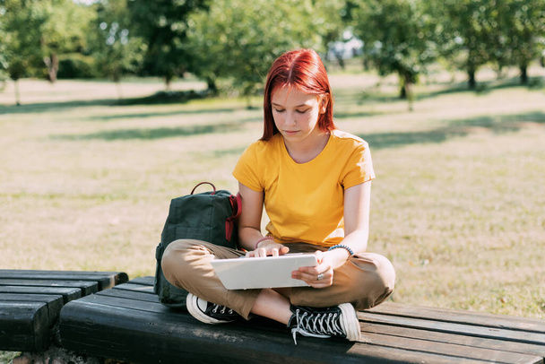 Genç bir kız elinde dizüstü bilgisayarla parkta bankta oturuyor ve derslere ya da sınavlara hazırlanıyor. Eğitim ve eğitim kavramı - Fotoğraf, Görsel