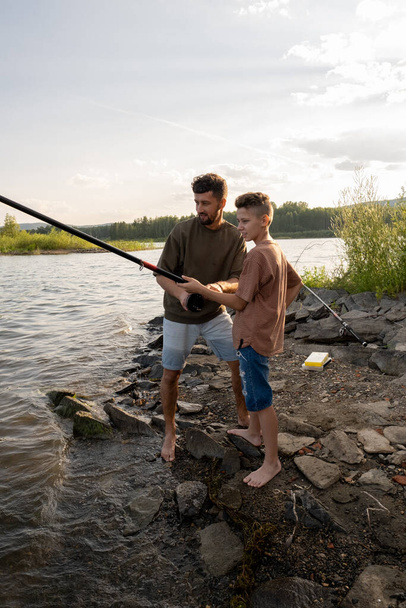 Молодой человек крутит колесо на удочке во время рыбалки со своим сыном - Фото, изображение