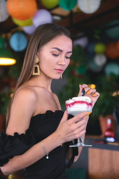 Beautiful sexy brunette woman in restaurant or cafe with ice cream tiramisu dessert. Fashion photo of young woman in black dress with hairstyle and makeup, eating dessert. Eat out concept. - 写真・画像