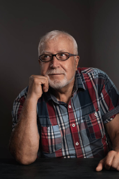 Elderly charismatic man in a checkered shirt, wearing glasses looks to the side with interest, close-up portrait on a gray background - Photo, Image