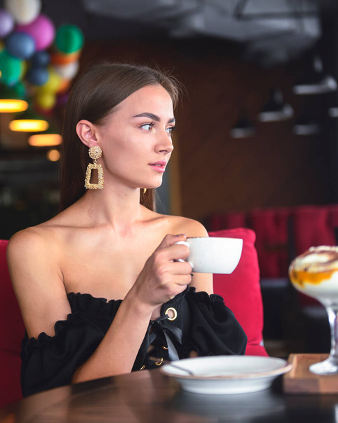 Beautiful sexy brunette woman in restaurant or cafe with ice cream tiramisu dessert and a cup of . Fashion photo of young woman in black dress with hairstyle and makeup eat out concept. - Photo, Image