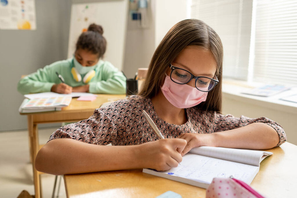 Χαριτωμένη επιμελής μαθήτρια με προστατευτική μάσκα κρατώντας σημειώσεις στο βιβλίο αντιγράφων - Φωτογραφία, εικόνα