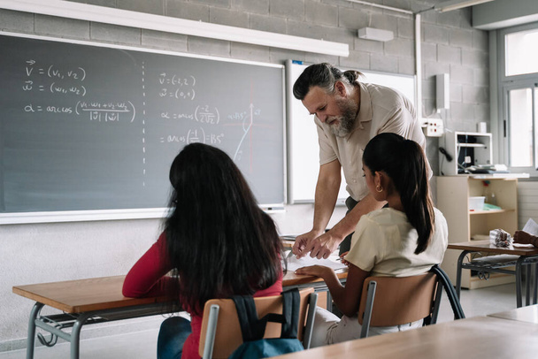Teacher with beard helping latin student in class. Professor explaining exam exercise to multi ethnic teenager students in college high school classroom - Photo, Image