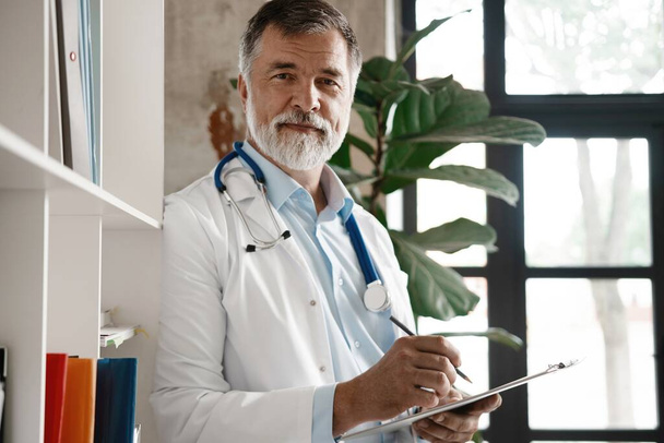 Portrait of handsome mature doctor standing at window in modern office. - Photo, Image