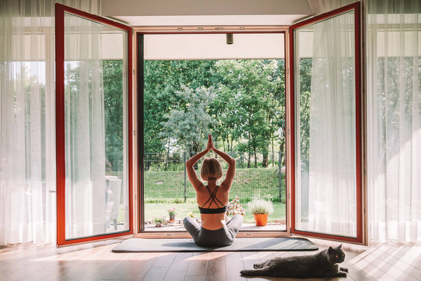 Woman sitting in a lotus position in front of the opened window, while doing yoga at home. Her cat is laying down next to her. - Фото, изображение