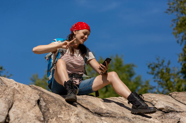 Hiker takes a rest and using mobile as a camera. Caucasian happy woman with a backpack and red bandana sitting on a rock and does selfie in sunny day in summer outdoor, selected focus. - Foto, Imagen