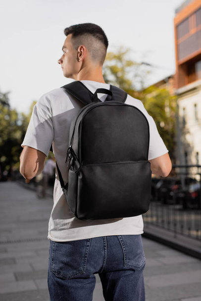 Rear view man walking in city with black leather backpack on his shoulders - 写真・画像