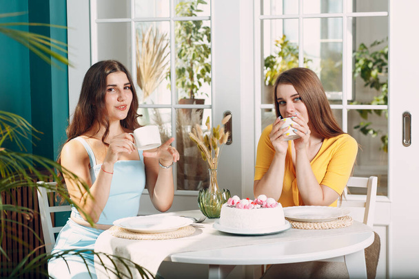 Mujeres amigas felices en casa sentadas y sonriendo con pastel de cumpleaños blanco y taza de té - Foto, imagen