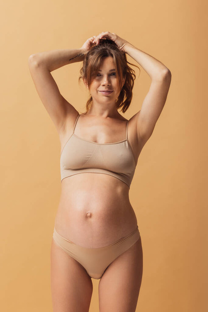 Portrait of young beautiful pregnant woman in lingerie isolated over sand color studio background. Natural beauty, happy motherhood, femininity concept. - 写真・画像