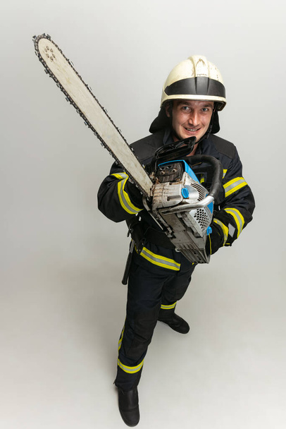 Jeden uśmiechnięty strażak ubrany w mundur pozował z piłą na białym tle studia. - Zdjęcie, obraz