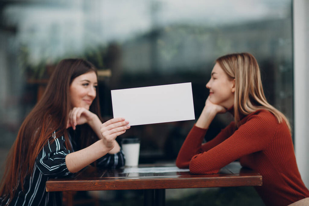 Woman holding white paper in hand. Two women talking at table in street cafe. White blank template sheet with empty space. - Photo, Image