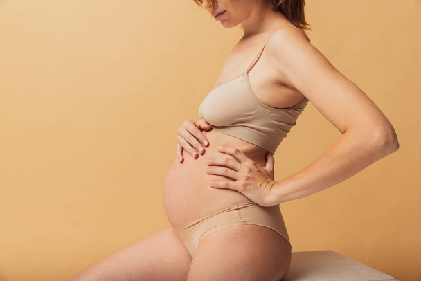 Portrait of young beautiful pregnant woman in lingerie isolated over sand color studio background. Natural beauty, happy motherhood, femininity concept. - 写真・画像