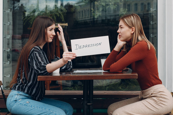 Γυναίκα κρατώντας λευκό χαρτί με ετικέτα λέξη κατάθλιψη στο χέρι. Δύο γυναίκες που μιλάνε στο καφέ του δρόμου. Έννοια κρυφής κατάθλιψης - Φωτογραφία, εικόνα