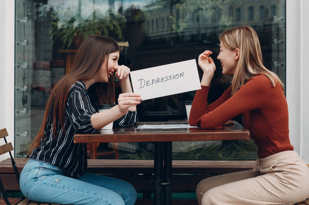 Piilotettu masennus käsite. Nainen, jolla on paperilappu, jossa lukee "Masennus kädessä". Kaksi naista puhuu ja hymyilee kahvilassa.. - Valokuva, kuva