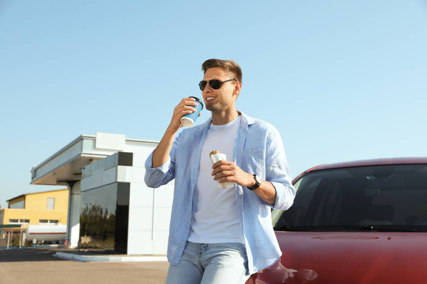 Young man with hot dog drinking coffee near car at gas station - Φωτογραφία, εικόνα