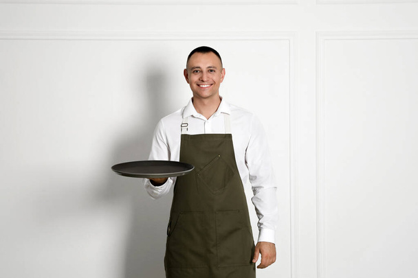 Portrait of happy young waiter with tray near white wall - Fotografie, Obrázek