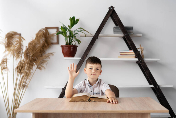 School boy thinks sitting at a desk in a home class with an open book learns waving hello to the camera. Pupil with a textbook does homework with a teacher in the zoom. Online education concept - Photo, Image