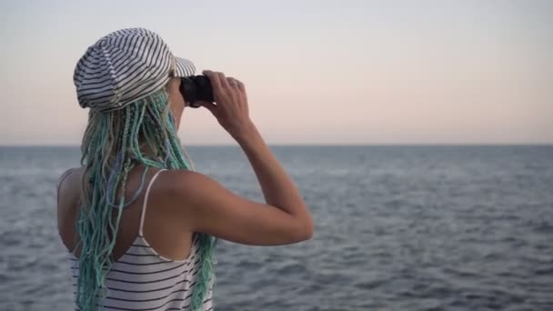 Een jonge vrouw in een marine gestreepte jurk kijkt door een verrekijker naar de zee of de oceaan - Video