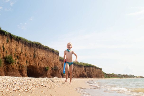 A boy runs along the beach in swimming trunks. He has a water pistol in his hands. Happy child on the sandy beach. Concept of happy life and relaxation by the ocean. Blond boy smiling at the camera - Photo, Image