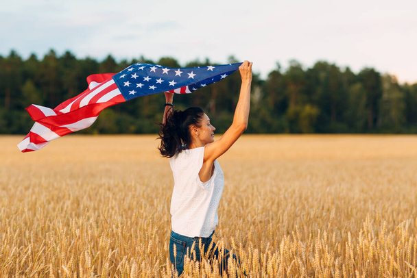 Nainen, jolla on Amerikan lippu vehnäpellolla auringonlaskun aikaan. Itsenäisyyspäivä. Itsenäisyyspäivä isänmaallinen loma. - Valokuva, kuva