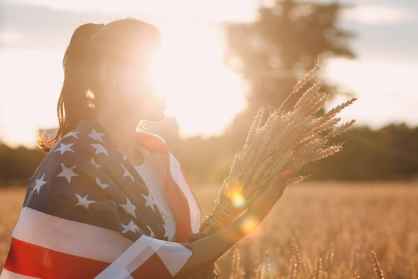 Woman with American flag and with a sheaf of ears in wheat field at sunset. 4th of July. Independence Day patriotic holiday. - 写真・画像