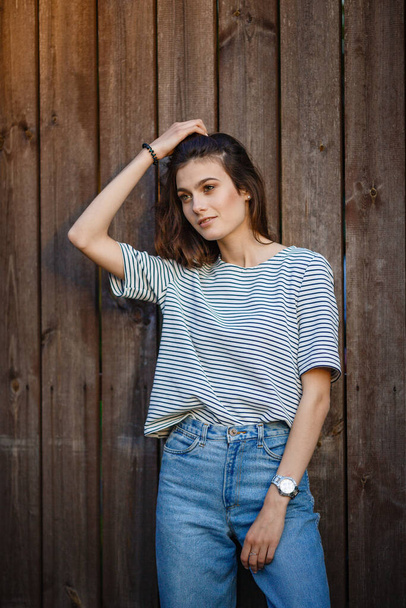 Portrait of a beautiful young brunette woman in striped sweater and high jeans against wall background of coarse-hewn gray boards. - Photo, Image