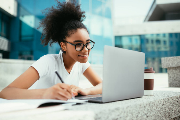 Onnellinen musta opiskelija tyttö tekee läksyjä käyttäen Laptop Outdoor - Valokuva, kuva