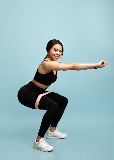 Strength workout concept. Full length of black woman doing squats with fitness resistance band over blue background - Photo, Image
