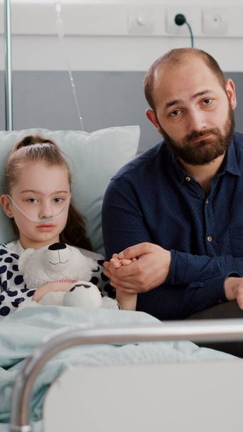 Endişeli baba portresi hastanede hasta kız elini tutuyor kameraya bakıyor. - Fotoğraf, Görsel