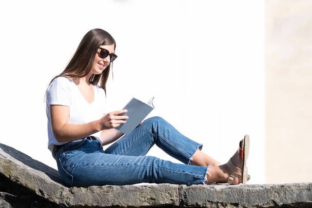 Młoda kobieta czytająca książkę na zewnątrz, siedząca na kamiennej ścianie. Styl życia studentów lub studentów uniwersytetów - Zdjęcie, obraz