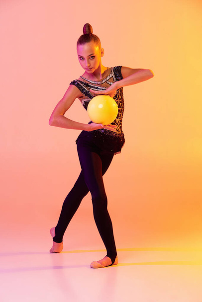 One young girl, rhythmic gymnastics artist in stage attire, sports costume isolated on pink yellow studio background in neon. Concept of sport - Foto, afbeelding