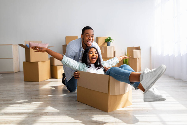 Moverse y concepto de bienes raíces. Alegre pareja afroamericana divirtiéndose y montando en cajas de cartón en nuevo apartamento - Foto, Imagen
