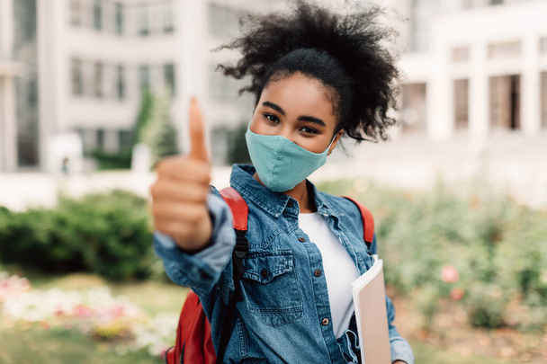 Fekete diák lány viselő arc maszk Gesturing hüvelykujj-up álló szabadban - Fotó, kép