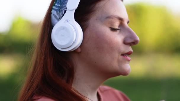 Mulher branca feliz vestindo fones de ouvido brancos ouvindo música com os olhos fechados na rua ao ar livre. - Filmagem, Vídeo