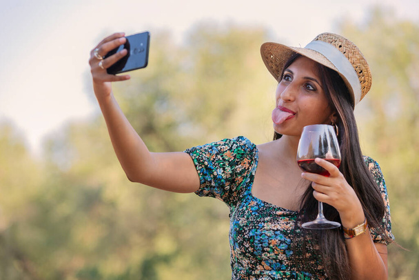 Funny fashionable young female with straw hat  shows tongue, makes selfie with mobile phone, holds wine glass in hands, enjoys recreation and good summer rest. Tourist woman in green park - Fotó, kép