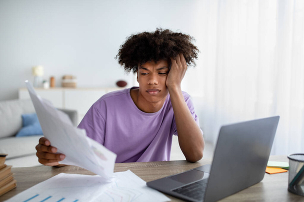 Набридлий підліток сидить за столом з документами, використовує ноутбук для написання курсового паперу, вивчає онлайн, готується до іспиту
 - Фото, зображення