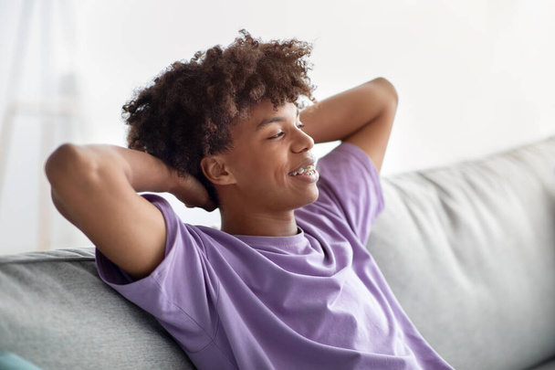 Tasasız zenci genç adam ellerini başının arkasına koyup kanepede dinleniyor ve tecrit sırasında evde vakit geçiriyor. - Fotoğraf, Görsel