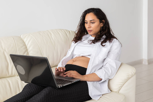 Mujer embarazada latina usando ordenador portátil sentado en el sofá en casa. Embarazo e información para el concepto de paternidad. - Foto, Imagen