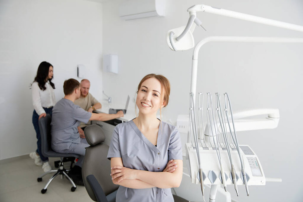 Молодая симпатичная женщина-дантист стоит в центре офиса, а на заднем плане коллега консультирует пациентов. Подготовка к приему пациентов - Фото, изображение