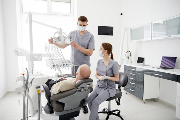 Sairaala kaksi lääkäriä lääketieteellisessä univormussa tutkii sairasta rauhallista suuta analysoimalla hampaiden terveyttä valmistautuen stomatologiseen leikkaukseen. Stomatologi lääkärit työskentelevät oikomishoidon klinikalla - Valokuva, kuva