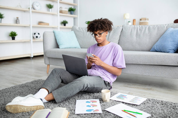 Teljes hossza fókuszált fekete tini srác használ okostelefon és laptop az online tanulás otthon - Fotó, kép