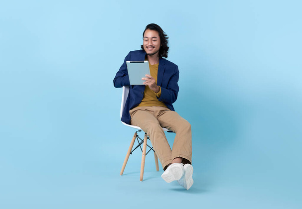 Businessman asian happy smiling using a digital tablet while sitting on chair isolated on bright blue background.	 - Foto, Imagen