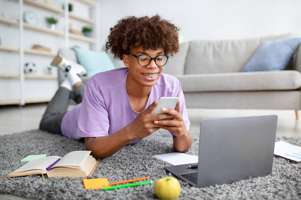 Happy black teen guy lying on floor with smartphone and laptop, studying online, checking social media at home - Photo, Image