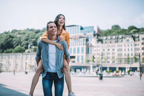 Happy young woman embracing and riding piggyback on joyful boyfriend while walking together on square during excited romantic summer tour  - Foto, afbeelding